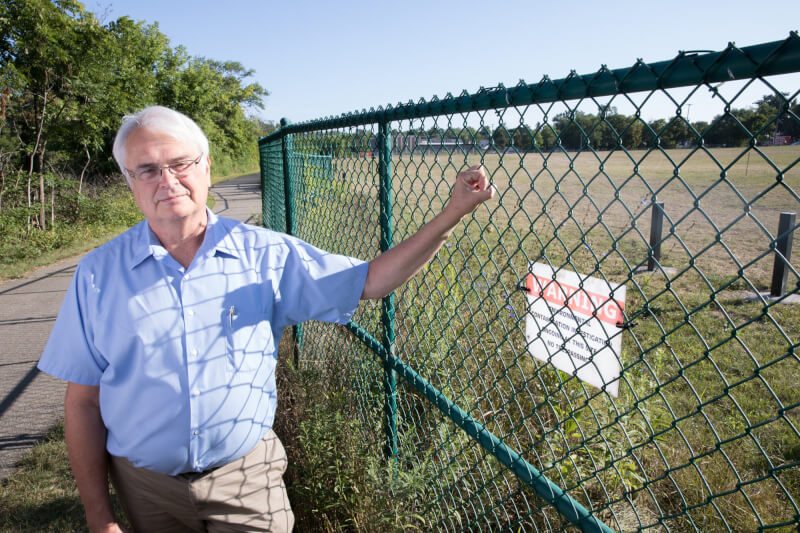 Rick Rediske stand next to the site of the former Wolverine World Wide tannery in Rockford, Michigan. He holds a chain link fence with a sign warning of environmental contamination.