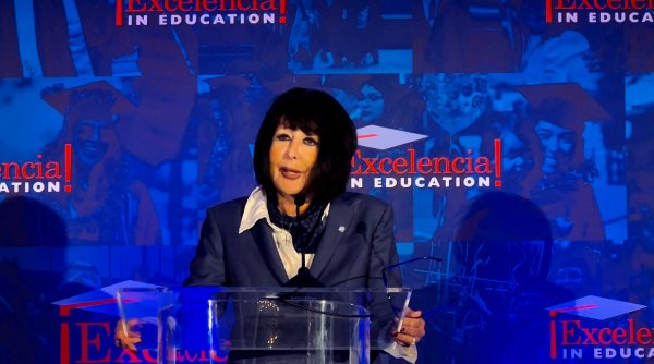President Mantella stands behind a podium speaking in front of a banner for Excelencia in Education