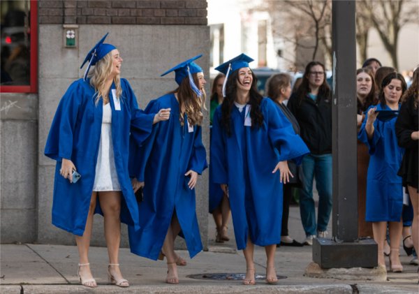Three people dressed in caps and gowns laugh on the curb before crossing the street. 