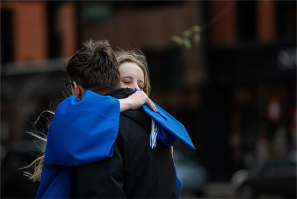  Two people in a heart-felt hug, one of them in a graduation gown holding their cap in their hand. 