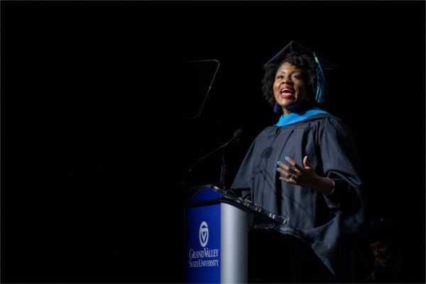 A person in a cap, gown, and hood stands at a podium speaking on stage. 