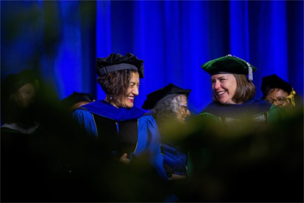 Two people sitting on stage wearing academic regalia smile and laugh together. 