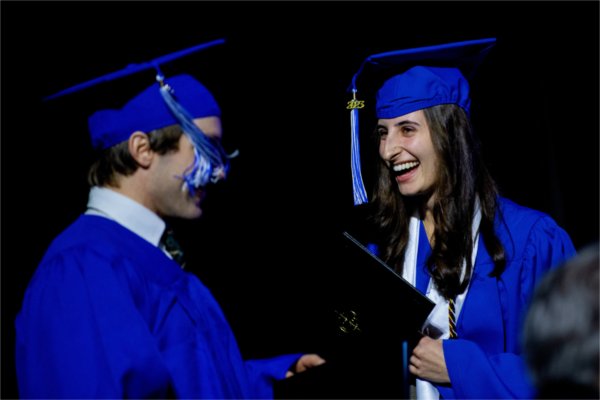 Two people in caps and gowns look at each other with big smiles while holding their degrees in hand. 