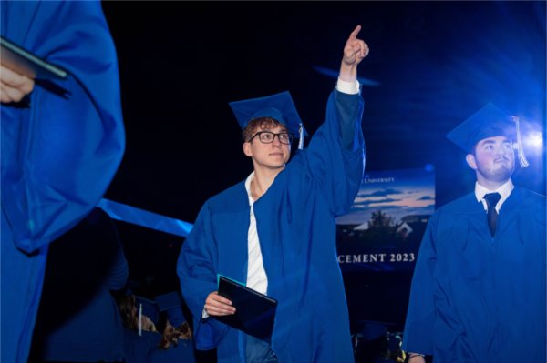 A person in a cap and gown holding their diploma holds their other hand up in the air pointing into the crowd.
