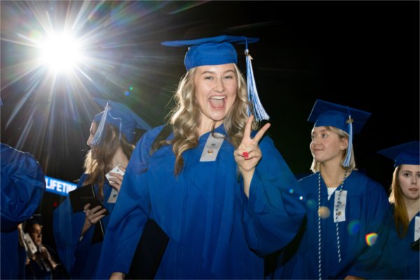 A person in a cap and gown holds out a peace sign and smiles at the camera. 