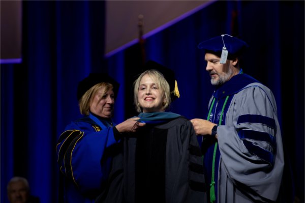 Three people in academic regalia stand on stage, two hood the person in the middle. 