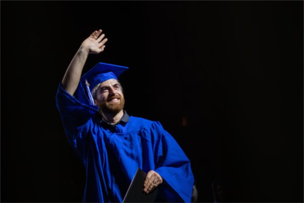 A person in cap and gown smiles and waves up into the crowd as they cross the stage. 