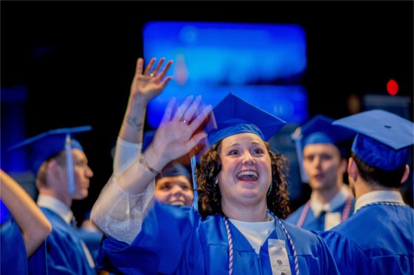 A person in cap and gown smiles and waves enthusiastically toward the crowd.