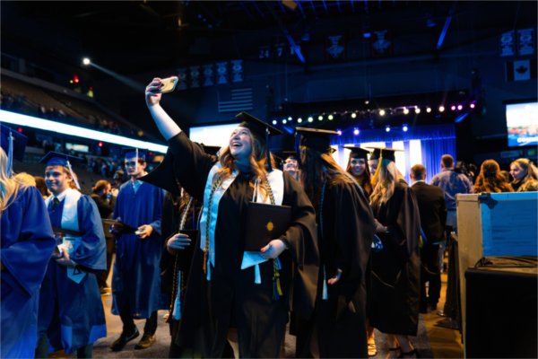 A person in a cap and gown holds their phone up high to get a selfie while exiting the arena. 