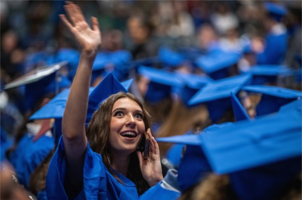 A soon-to-be graduate waves into the crowd while holding their cell phone to their ear and surrounded by other soon-to-be graduates.