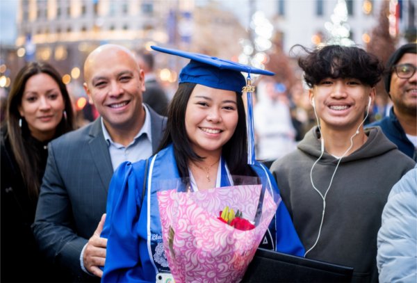 A person in cap and gown holds flowers while smiling and looking at the camera with their family around them. 