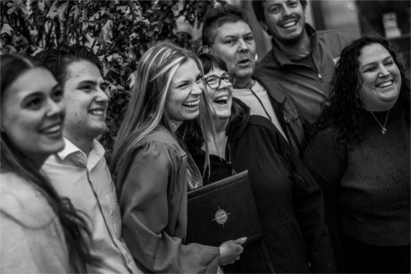 A recent graduate surrounded by people, all smiling and looking at a camera out of frame. 