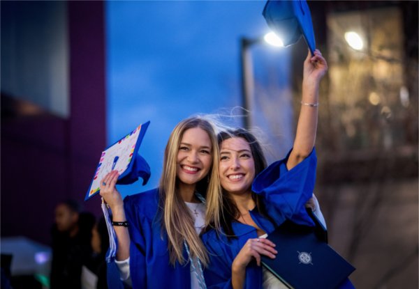 Two people stand outside in their graduation gowns while holding their caps in the air after Commencement. 