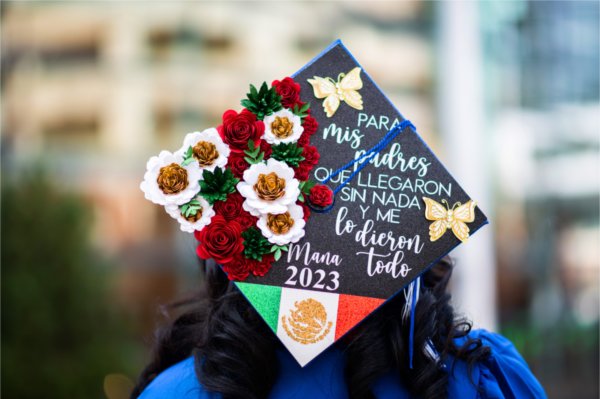 A graduate stands with their back to the camera showing off the top off their cap decorated with white and red flowers, a flag, and butterflies and the words read in Spanish, "My parents came with nothing to give it all." 