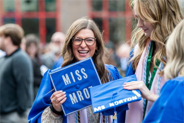 Three people in graduation gowns stand close to each other holding their caps in their hands. One faces the camera directly and her cap reads, "Miss Mroz".