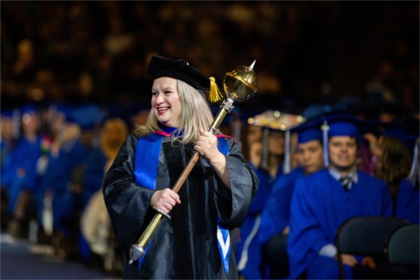 A person in academic regalia, holding a ceremonial mace, proceeds into the arena toward the stage while soon-to-be graduates look on. 