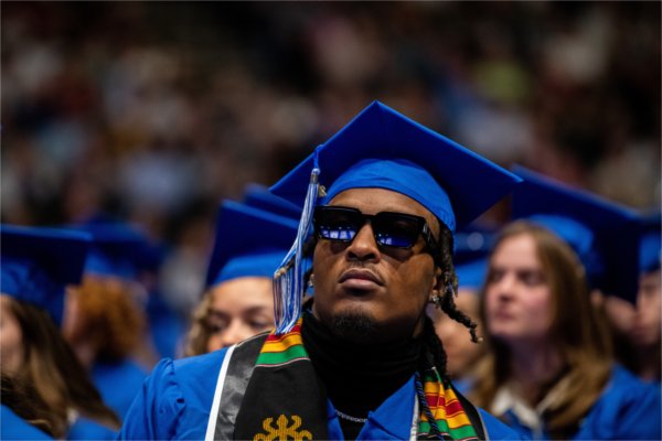 A person dressed in a cap and gown and wearing sunglasses sits in a crowd of soon-to-be graduates. 