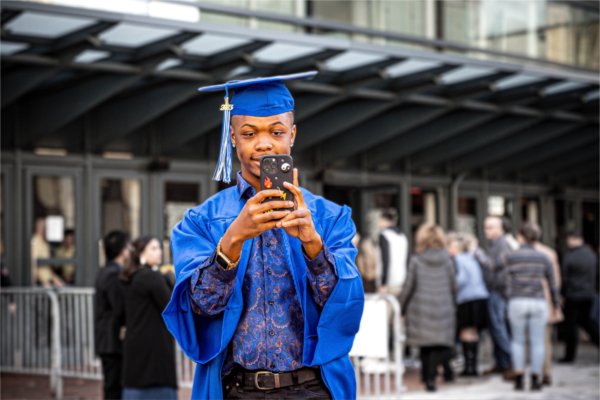 A soon-to-be graduate poses for a selfie in their cap and gown in front of a building. 
