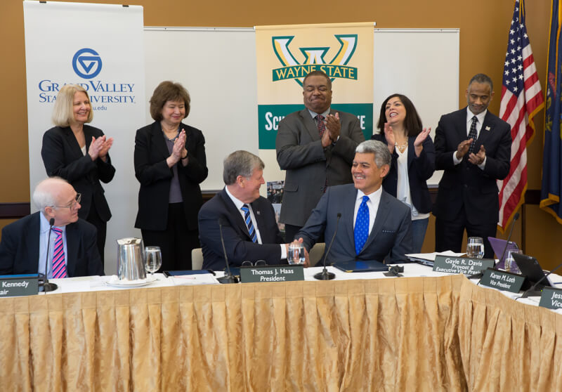 Two men shake hands in front of row of people who are standing