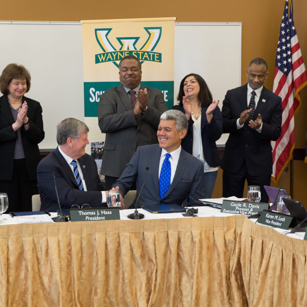 Two men shake hands in front of row of people who are standing