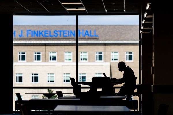  A student working on their laptop is silhouetted against a window in an academic building. 
