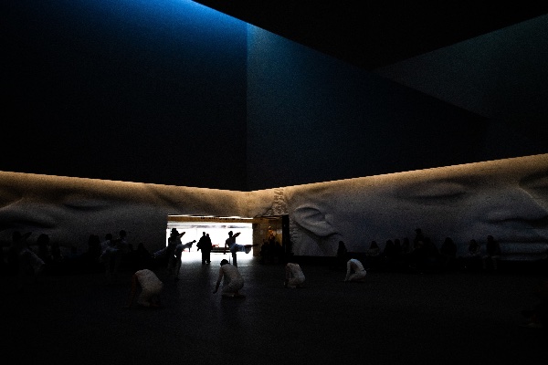 Dancers are silhouetted in an entrance to a room with face sculptures on the wall. 