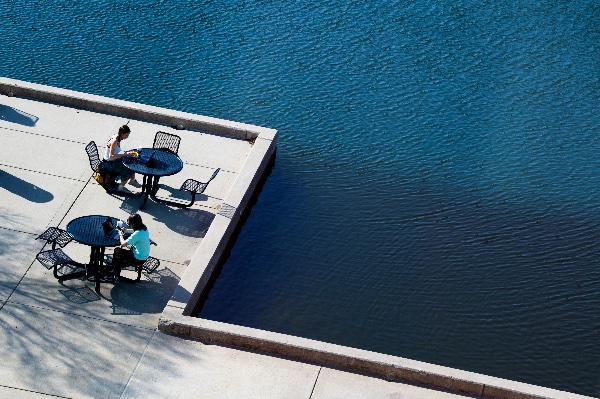  As seen from above, students sit at tables enjoying lunch along a blue pond. 