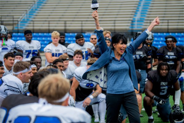 President Philomena V. Mantella holds a microphone and raises both hands in excitement while surrounded by the GVSU football team.
