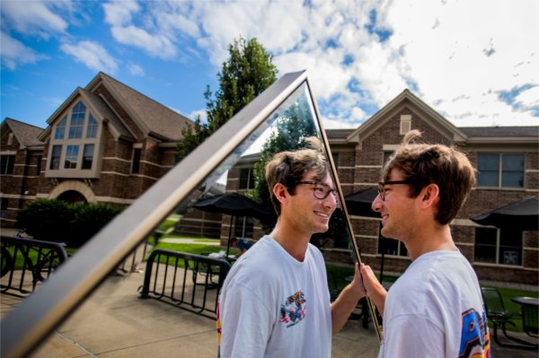 A person wearing a white graphic tee smiles while holding a mirror towards himself.