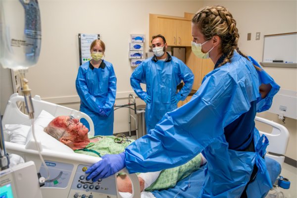 A student with braided hair wearing protective gear stands with two others as they examine a patient laying on a hospital bed.
