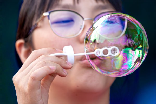  A close-up of person with glasses blowing a bubble towards the camera.