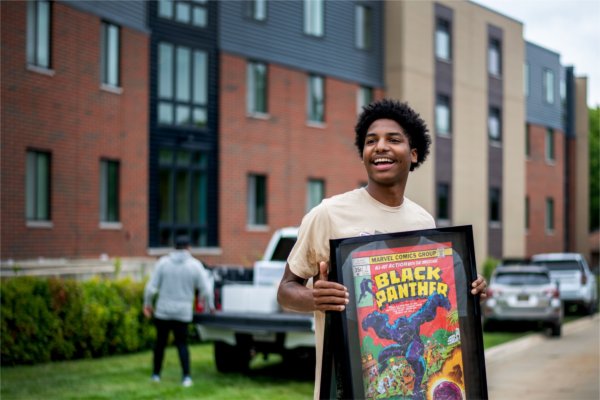 A person smiles holding up a framed picture of Black Panther with dorm buildings in the background.