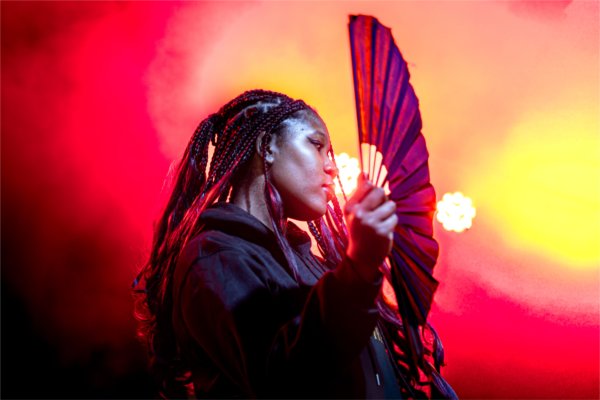  A side-view of a person with braids holding a handheld fan open. Bright red and yellow lights illuminate the background.