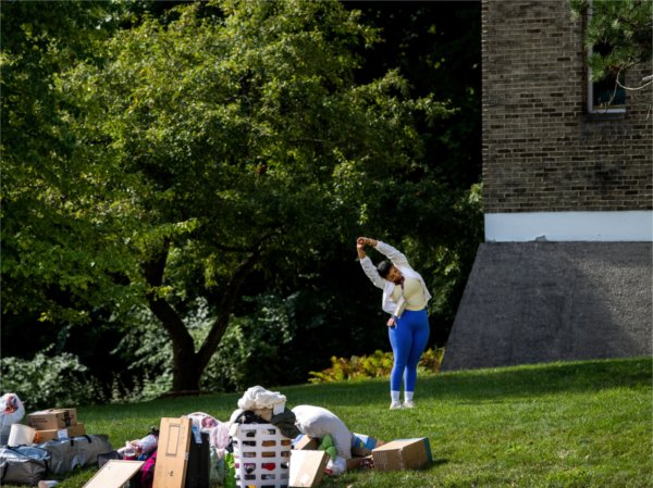  A person with bright blue leggings holds her hands together as stretches out her arms on the grass. Piles of move-in items are set on the grass in front of her.