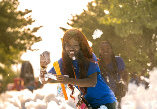 A person with dark red hair wearing a blue shirt laughs while standing in a foam pit.