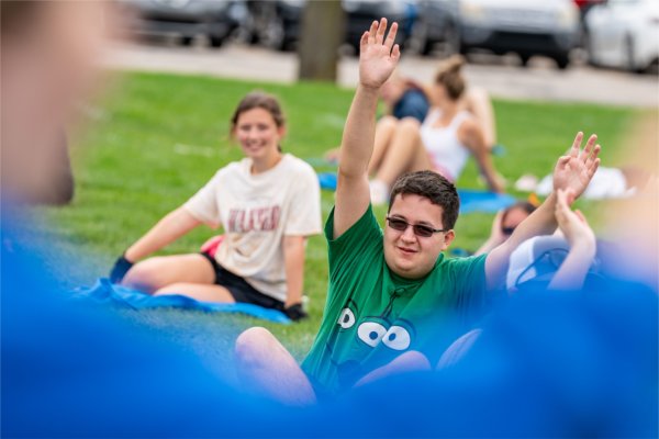 A person in shaded glasses wearing a green shirt raises both arms in the air as they sit on the grass.