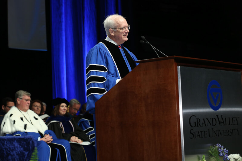 man on stage in academic robe, speaking; people seated in chairs behind him