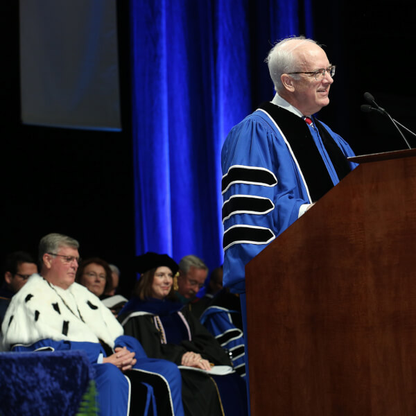 man on stage in academic robe, speaking; people seated in chairs behind him