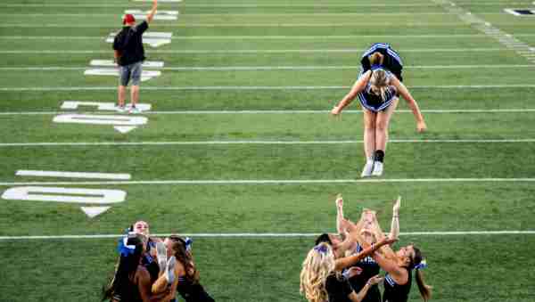 A cheerleader is tossed into the air along the sidelines of a football field.  
