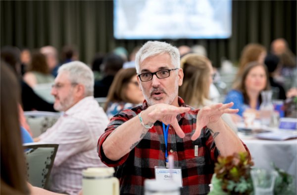 Chad Frederick in plaid shirt gesturing with hands while seated at table. Others fill in at tables around him
