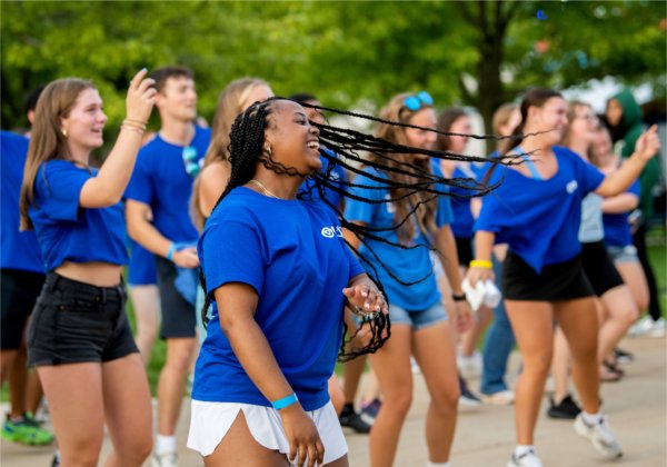 A young woman with long braided hair dances with a group of other people, all wearing blue t-shirts. 