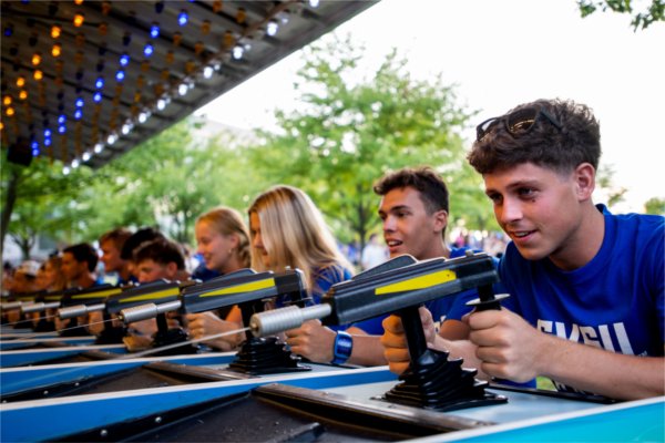 A line of people in blue t-shirts play a water gun shooting carnival game. 