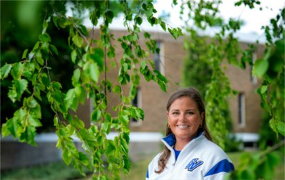 Colleen Bailey wears a white and blue GV quarter zip and stands beneath a tree in front of Kistler Living Center. 