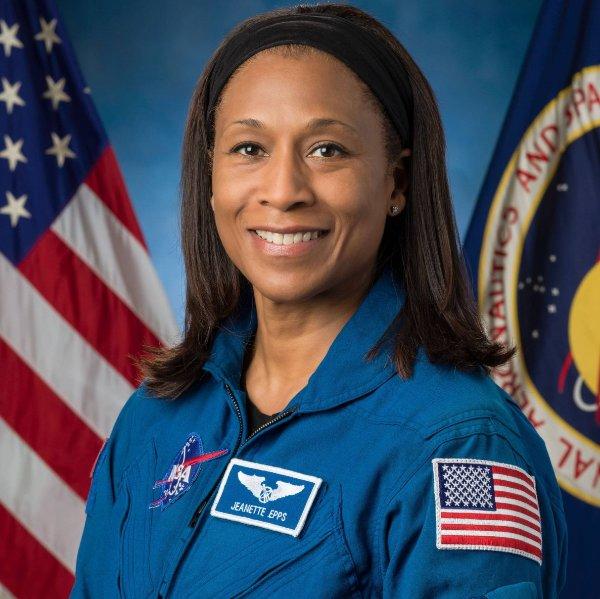 A person wearing an astronaut uniform smiles for a portrait. An American flag is in the background.