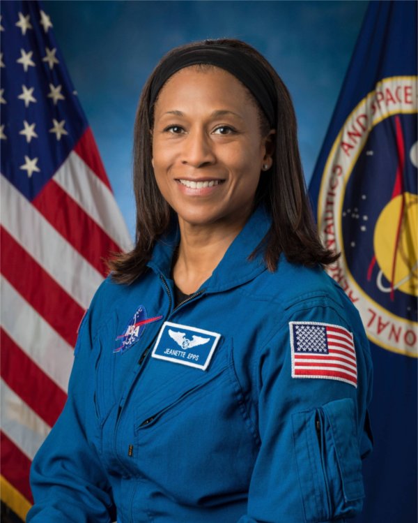 A person wearing an astronaut uniform smiles for a portrait. An American flag is in the background.