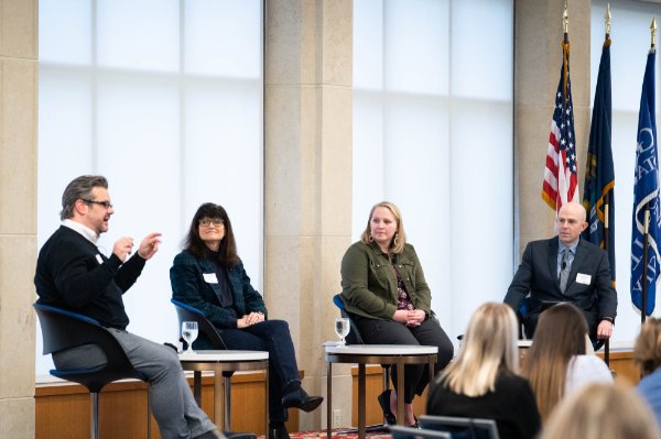 four people seated on stage, man on left gestering with hands