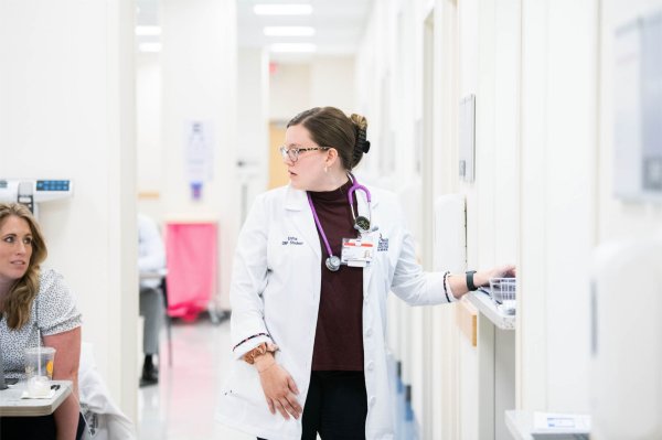 woman in white lab coat next to a door, long hallway of doors visible, woman seated at left