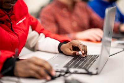 Close-up of a person in a red jacket using a laptop in a classroom, with other students working on computers in the background.