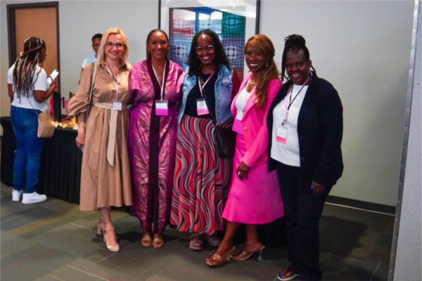 Five women wearing name badges pose together at an event, smiling at the camera. They stand indoors near a registration table and colorful abstract artwork.