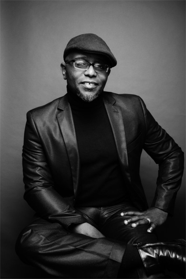 A person wearing a hat and glasses, sitting with one leg crossed over the other, smiles for the camera.